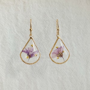 May include: A pair of gold teardrop-shaped earrings with a delicate purple flower pressed inside each earring.