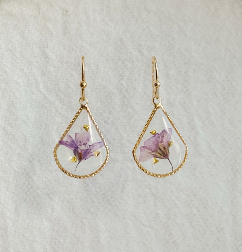 May include: A pair of gold teardrop-shaped earrings with a delicate purple flower pressed inside each earring.