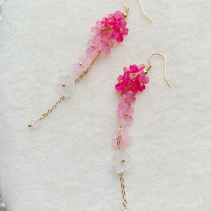 Puede incluir: Un par de pendientes largos y delicados con un degradado de flores rosas y blancas que cuelgan de cadenas doradas.