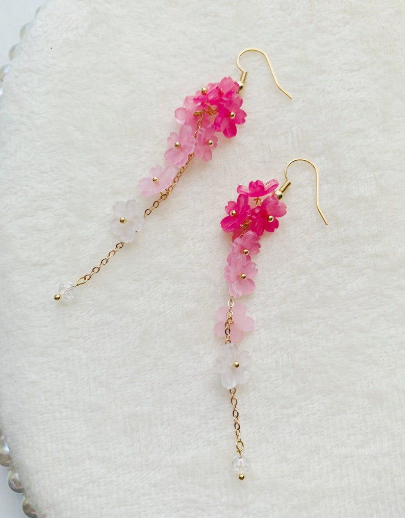 Puede incluir: Un par de pendientes largos y delicados con un degradado de flores rosas y blancas que cuelgan de cadenas doradas.