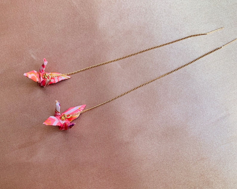 Może przedstawiać: Para długich, złotych kolczyk&oacute;w z wiszącymi origami z papierowych żurawi. Żurawie są r&oacute;żowe, białe i pomarańczowe z kwiatowym wzorem.