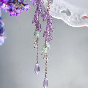 Può includere: Orecchini pendenti dal design floreale. Gli orecchini presentano grappoli di piccole perline viola chiaro, intervallate da perline verde pallido e dettagli color argento. Ogni orecchino termina con un cristallo a forma di goccia, viola chiaro.