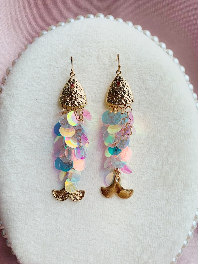 May include: A pair of dangle earrings with a mermaid theme. The earrings feature gold-colored textured triangular tops with a small red gem, iridescent sequin strands, and a gold-colored mermaid tail charm.