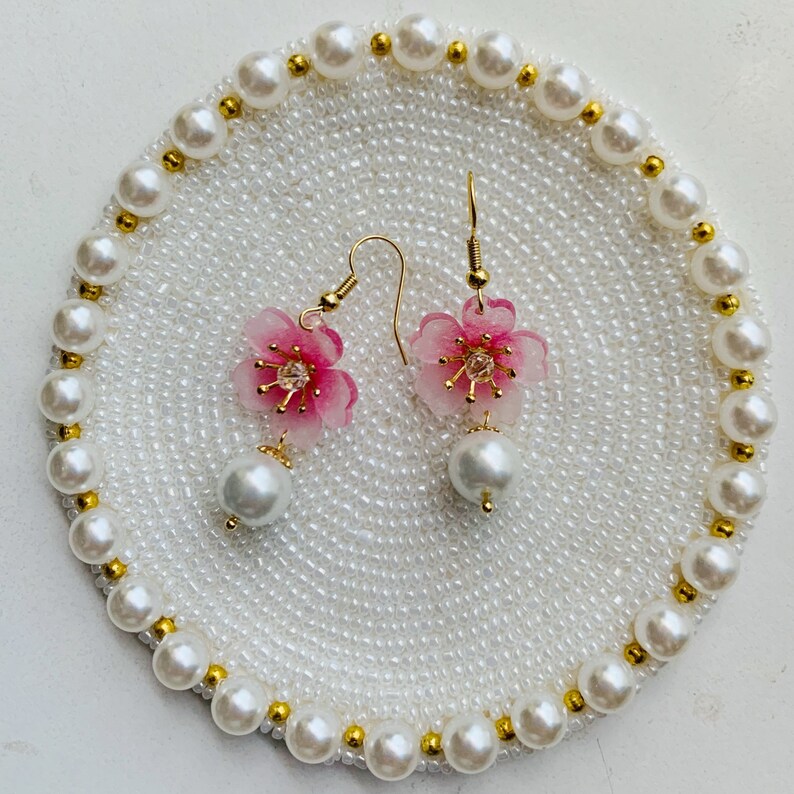May include: A pair of pink and white flower earrings with gold accents and pearl drops. The earrings are displayed on a white pearl-covered dish.