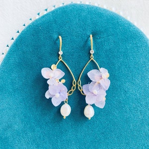May include: Gold-tone drop earrings with lavender fabric flower clusters and a dangling white pearl. The earrings have a teardrop shape with gold filigree and small clear crystals. Each earring is approximately 6 cm long.