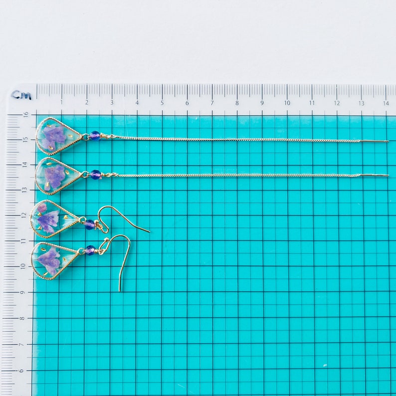 May include: Three pairs of dangle earrings with purple pressed flowers encased in clear resin. The earrings are on a white surface with a blue grid.
