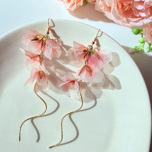 May include: A pair of delicate, floral drop earrings. Each earring features cascading pink fabric flowers with gold-colored accents and a long, wavy gold-tone chain. The earrings are displayed on a white plate, with pink flowers in the background.