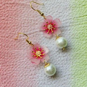 May include: A pair of dangle earrings featuring pink cherry blossom flower charms with gold accents and white pearl drops.