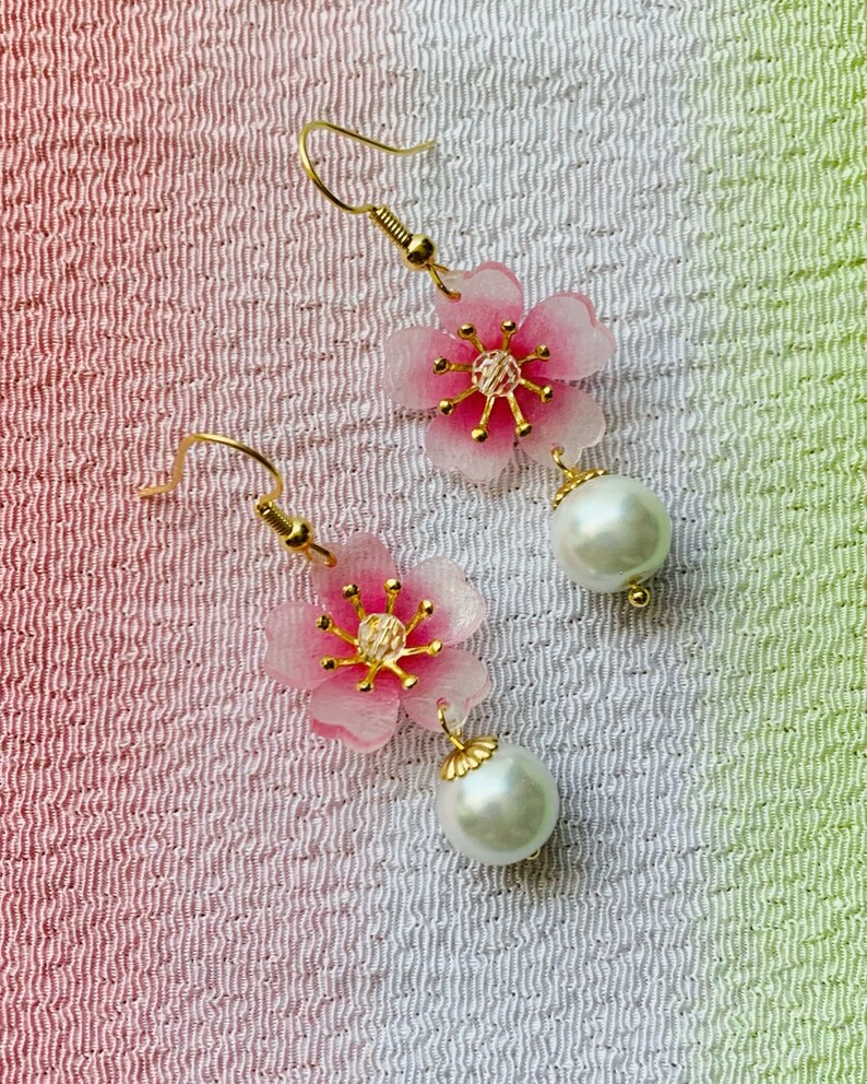May include: A pair of dangle earrings featuring pink cherry blossom flower charms with gold accents and white pearl drops.