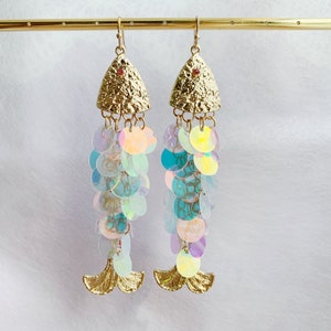 May include: A pair of gold-colored fish-shaped earrings. The earrings feature textured gold tops with a small red gem and iridescent sequin strands. The bottom of the earrings have gold-colored fin details.