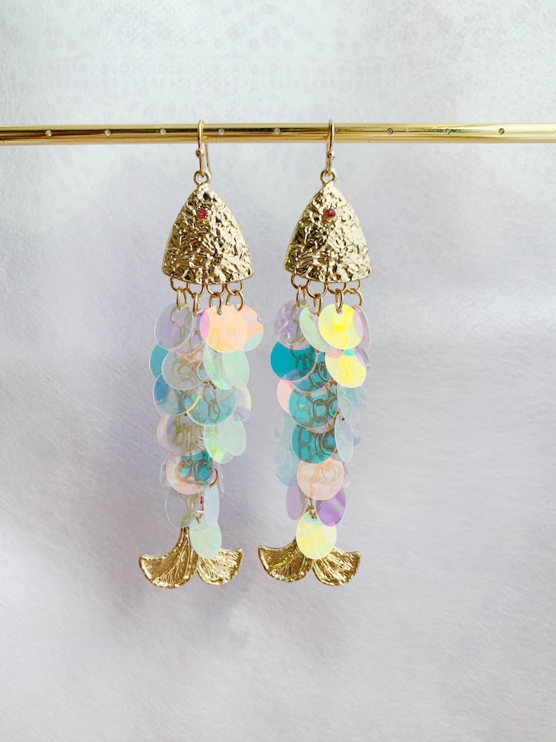 May include: A pair of gold-colored fish-shaped earrings. The earrings feature textured gold tops with a small red gem and iridescent sequin strands. The bottom of the earrings have gold-colored fin details.
