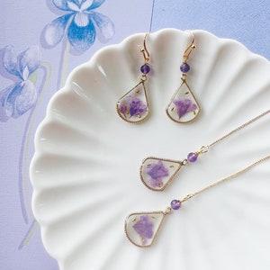 May include: A set of delicate gold earrings and a necklace featuring teardrop-shaped pendants with pressed purple flowers inside. The earrings have amethyst-colored beads at the top of the teardrop.