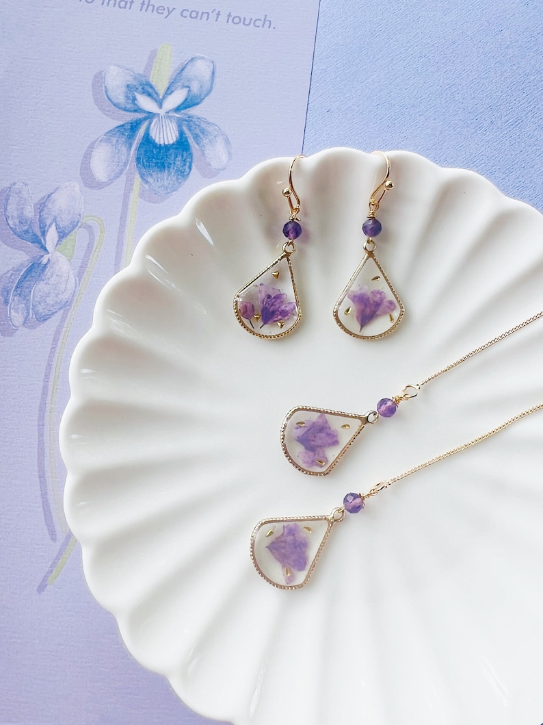 May include: A set of delicate gold earrings and a necklace featuring teardrop-shaped pendants with pressed purple flowers inside. The earrings have amethyst-colored beads at the top of the teardrop.