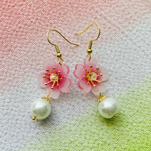May include: A pair of gold earrings featuring pink flower charms and white pearl accents. The flowers have a translucent, almost glass-like appearance.
