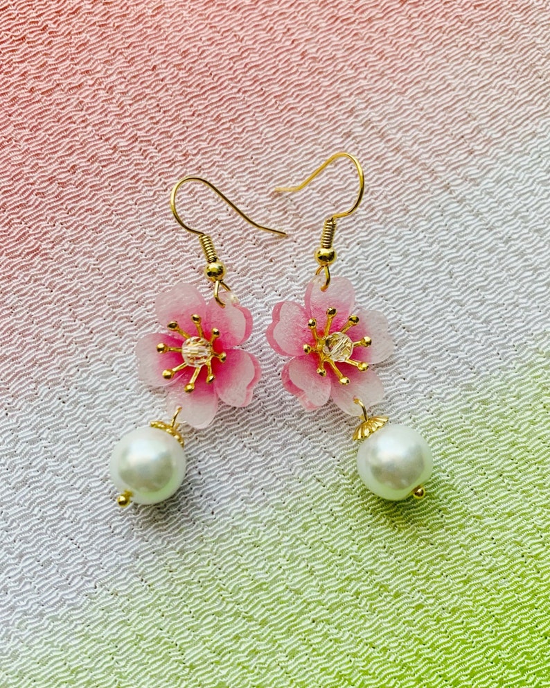 May include: A pair of gold earrings featuring pink flower charms and white pearl accents. The flowers have a translucent, almost glass-like appearance.