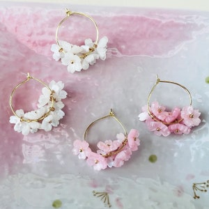 May include: Four pairs of gold hoop earrings adorned with clusters of white and pink cherry blossom-shaped beads. The earrings are arranged on a pink and white surface, creating a delicate and feminine aesthetic.