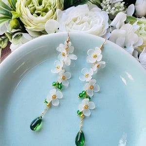 Può includere: Un paio di orecchini pendenti con ciondoli a forma di fiore bianco e cristalli verdi a goccia.