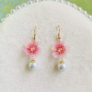 May include: A pair of pink cherry blossom earrings with gold accents and a pearl dangle. The earrings are on a white background.