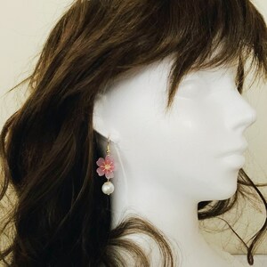 May include: A pair of dangle earrings featuring a pink flower charm and a white pearl bead. The earrings are displayed on a white mannequin head with brown hair.