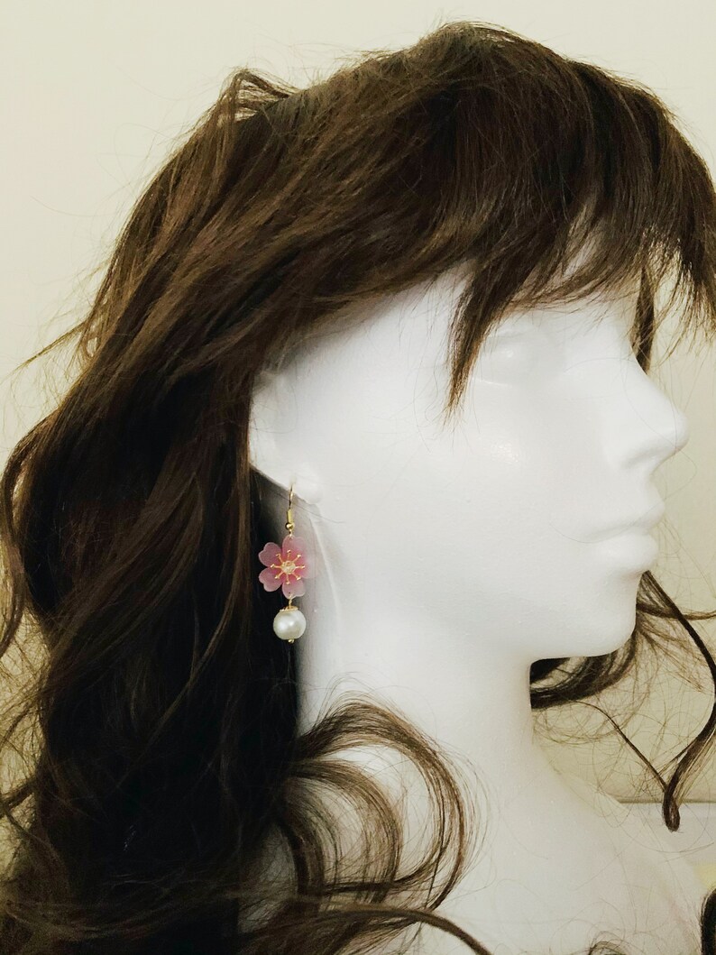 May include: A pair of dangle earrings featuring a pink flower charm and a white pearl bead. The earrings are displayed on a white mannequin head with brown hair.
