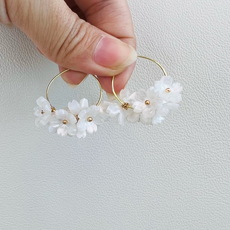 May include: Gold hoop earrings adorned with delicate white flower clusters. Each earring features multiple small, petal-like shapes and tiny gold accents, creating a floral design. The earrings are held against a white background.