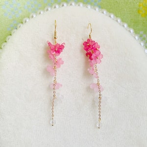 Puede incluir: Un par de pendientes colgantes con un dise&ntilde;o de flores rosa y blancas. Los pendientes est&aacute;n hechos con peque&ntilde;as flores delicadas que est&aacute;n enhebradas en una cadena dorada. Los pendientes est&aacute;n terminados con una cuenta de cristal transparente en la parte inferior.