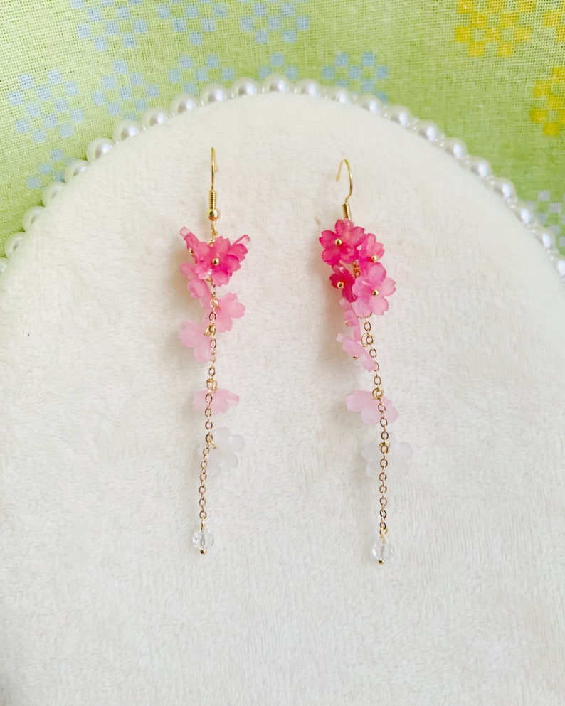 Puede incluir: Un par de pendientes colgantes con un dise&ntilde;o de flores rosa y blancas. Los pendientes est&aacute;n hechos con peque&ntilde;as flores delicadas que est&aacute;n enhebradas en una cadena dorada. Los pendientes est&aacute;n terminados con una cuenta de cristal transparente en la parte inferior.