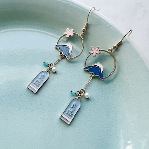May include: A pair of gold earrings with a blue and white mountain design, pink flowers, and a charm with Japanese characters. The earrings are hanging from a blue plate.