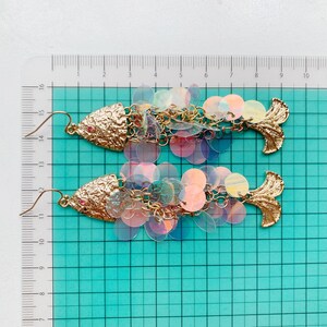 May include: A pair of gold-toned fish-shaped earrings. The earrings feature textured gold heads with a small red accent and a tail fin. The fish bodies are made of iridescent, round sequins, connected by gold chains.