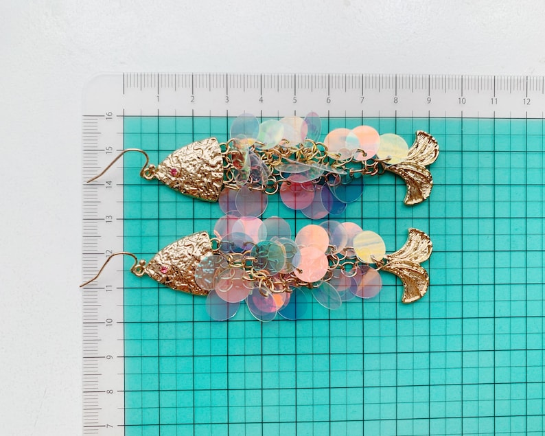 May include: A pair of gold-toned fish-shaped earrings. The earrings feature textured gold heads with a small red accent and a tail fin. The fish bodies are made of iridescent, round sequins, connected by gold chains.