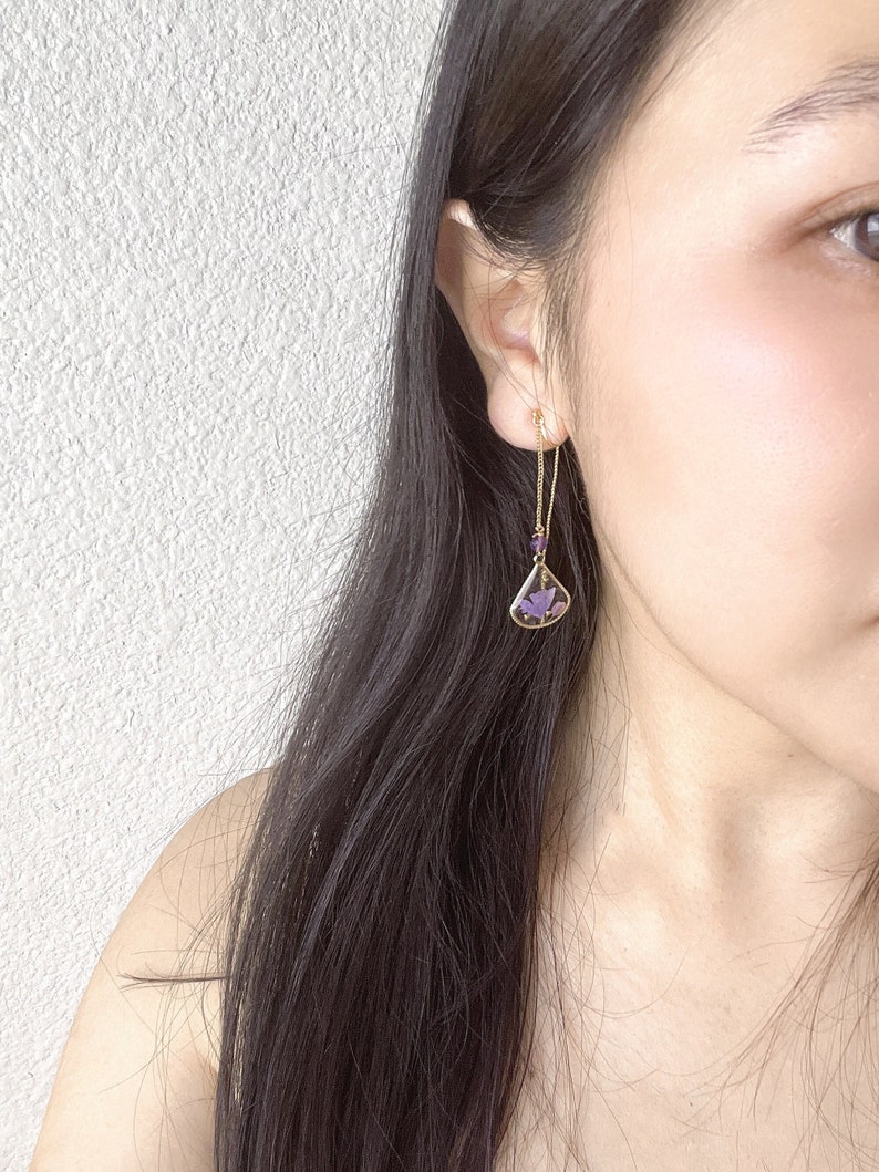 May include: A pair of gold dangle earrings with a purple butterfly design. The earrings are made of a delicate chain with small purple beads.