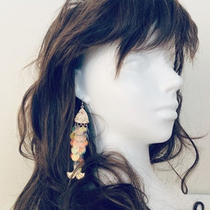 May include: A pair of dangle earrings with a fish design. The earrings feature iridescent, colorful sequins and gold-colored metal accents. The earrings are displayed on a white mannequin head with brown hair.