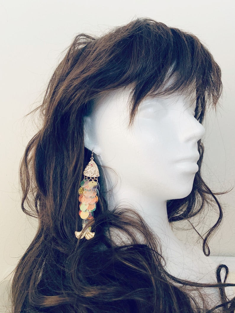 May include: A pair of dangle earrings with a fish design. The earrings feature iridescent, colorful sequins and gold-colored metal accents. The earrings are displayed on a white mannequin head with brown hair.