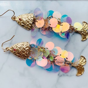 May include: A pair of gold-colored fish-shaped earrings. The head of each earring has a textured finish with a small red gem. The body is made of iridescent, round sequins in shades of pink, blue, and yellow. The tail is also gold-colored.