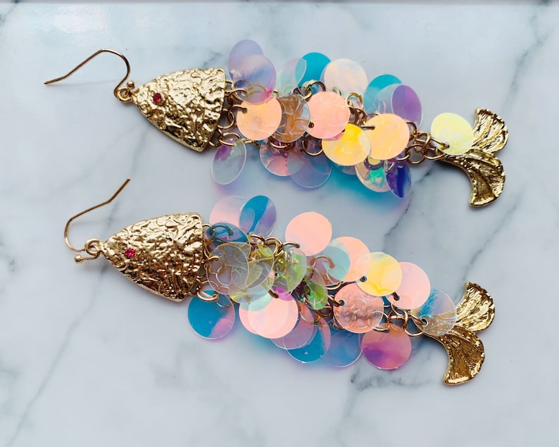 May include: A pair of gold-colored fish-shaped earrings. The head of each earring has a textured finish with a small red gem. The body is made of iridescent, round sequins in shades of pink, blue, and yellow. The tail is also gold-colored.