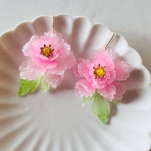Könnte beinhalten: Ein Paar rosa Pfingstrosen-Ohrringe mit goldfarbenen Haken. Die Ohrringe haben ein durchscheinendes Aussehen mit grünen Blättern und gelben Zentren. Die Ohrringe werden auf einem weißen, wellenförmigen Teller präsentiert.