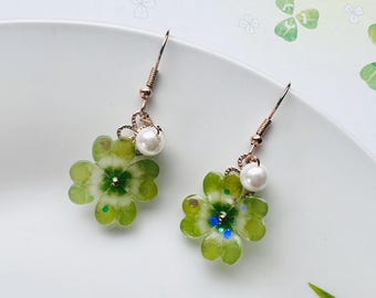 Peut inclure: Boucles d'oreilles faites à la main avec des motifs de trèfles à quatre feuilles. Chaque boucle d'oreille a un trèfle vert clair translucide avec un accent de perle et des crochets dorés. Les boucles d'oreilles sont présentées sur une assiette blanche avec un fond sur le thème du trèfle.