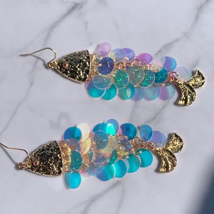 May include: A pair of fish-shaped earrings. The earrings feature a gold-colored textured head and tail, with iridescent sequins in shades of blue, green, and purple forming the body. The earrings have gold-colored ear wires.