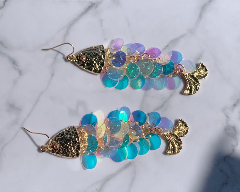 May include: A pair of fish-shaped earrings. The earrings feature a gold-colored textured head and tail, with iridescent sequins in shades of blue, green, and purple forming the body. The earrings have gold-colored ear wires.