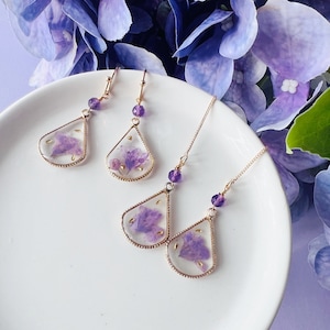 May include: A set of three pairs of earrings featuring delicate gold-toned teardrop-shaped pendants with pressed purple flowers inside. The earrings are displayed on a white plate.