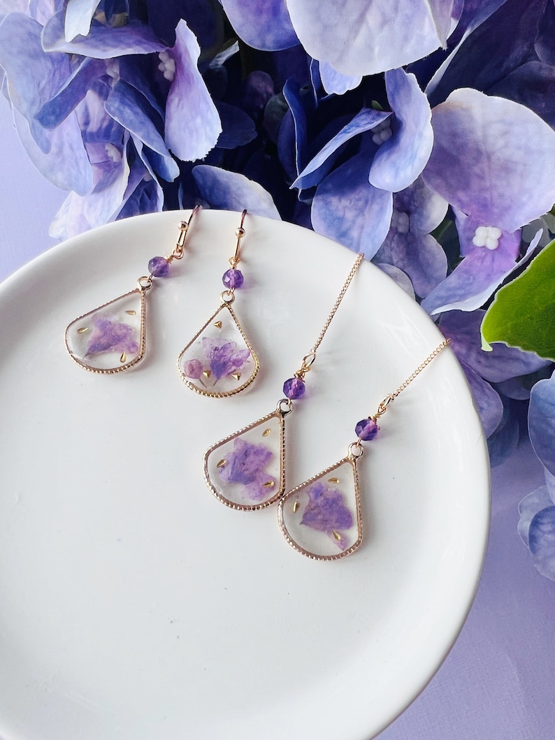 May include: A set of three pairs of earrings featuring delicate gold-toned teardrop-shaped pendants with pressed purple flowers inside. The earrings are displayed on a white plate.