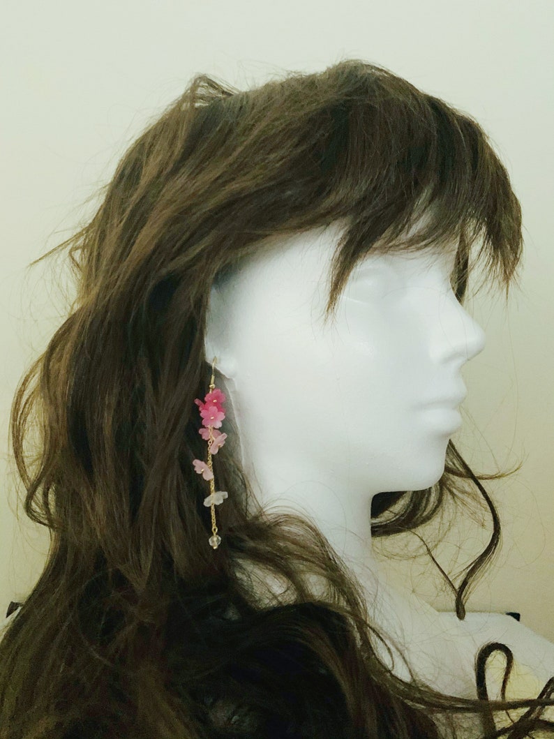 Puede incluir: Un par de pendientes largos y colgantes con una cadena dorada y abalorios en forma de flores rosas y blancas.