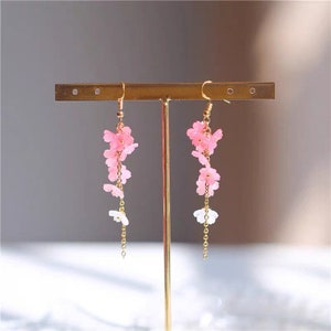 Puede incluir: Un par de aretes largos y delicados con dijes de flores rosas y blancas que cuelgan de cadenas doradas.