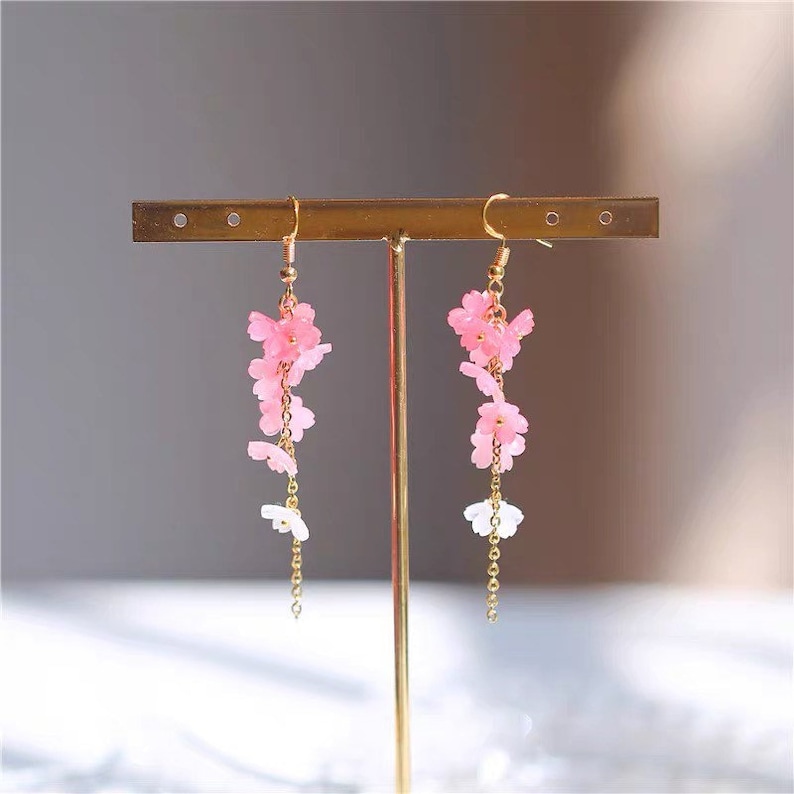 Puede incluir: Un par de aretes largos y delicados con dijes de flores rosas y blancas que cuelgan de cadenas doradas.