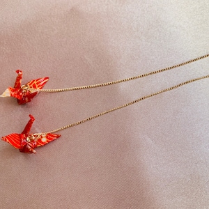 Może przedstawiać: Para czerwonych kolczyk&oacute;w z origami w kształcie żurawia z złotymi łańcuszkami. Żurawie są złożone z wzorzystego papieru i mają delikatny, skomplikowany wz&oacute;r.