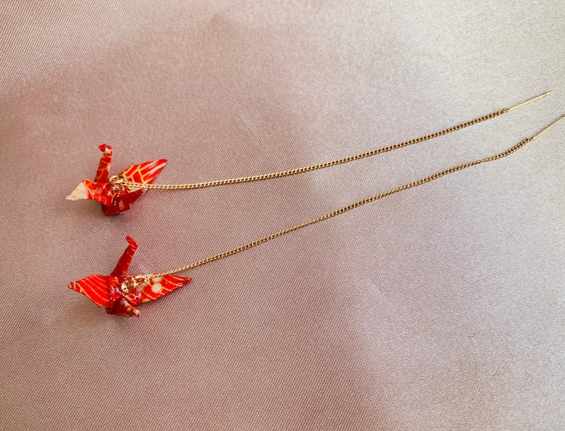 Może przedstawiać: Para czerwonych kolczyk&oacute;w z origami w kształcie żurawia z złotymi łańcuszkami. Żurawie są złożone z wzorzystego papieru i mają delikatny, skomplikowany wz&oacute;r.