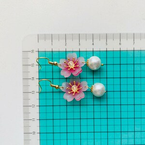 May include: A pair of pink and white flower earrings with gold accents and pearl drops. The earrings are approximately 3.8 cm long.