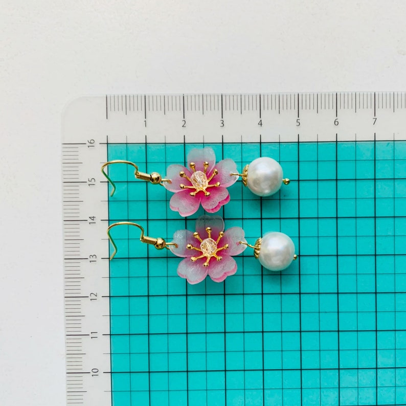 May include: A pair of pink and white flower earrings with gold accents and pearl drops. The earrings are approximately 3.8 cm long.