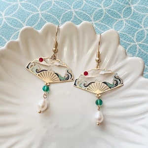 May include: Gold earrings with a fan design featuring a white and blue seascape with a red sun. The earrings have a green bead and a pearl drop.
