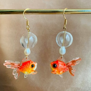 Peut inclure: Boucles d'oreilles représentant des poissons rouges. Chaque boucle d'oreille est ornée d'un petit poisson orange et jaune avec des nageoires transparentes, une perle transparente, une perle blanche et un crochet doré.